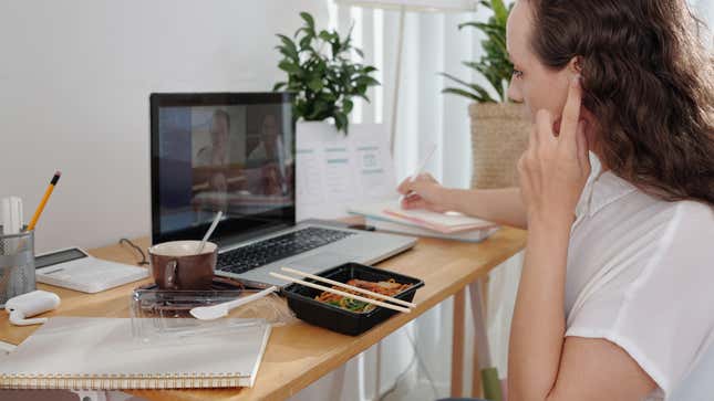 Bir kadın öğle yemeği yerken ve not defterine notlar alırken bir dizüstü bilgisayarda çalışır.